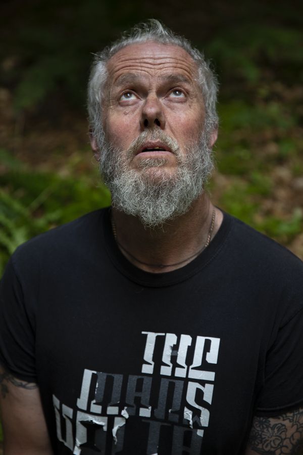 Bearded man with tattoos looks upward, wearing a black shirt outdoors.