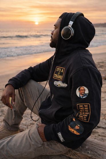 Man in headphones sitting on beach at sunset, wearing a hoodie with patches.