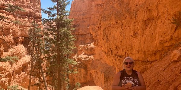 Woman hiking joyfully on a rocky trail surrounded by tall orange cliffs and pine trees.