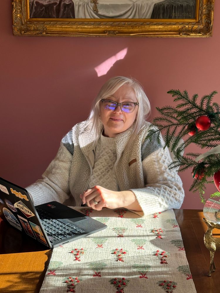A woman with white hair and glasses working on a laptop at a decorated wooden table.