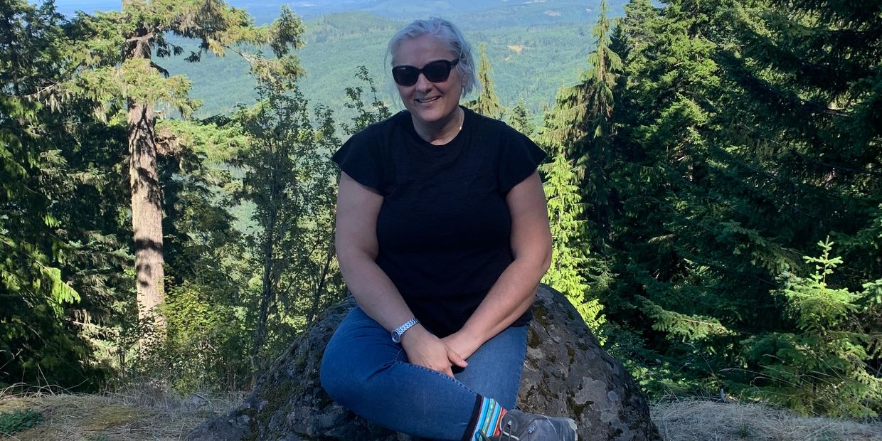 Woman in sunglasses sits on a rock with a scenic mountain and forest view.