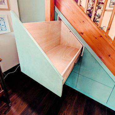 Custom-built pull-out storage drawer under stairs with angled design.