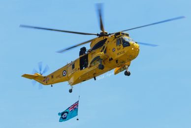 Yellow rescue helicopter flying with a flag beneath it against a clear blue sky.