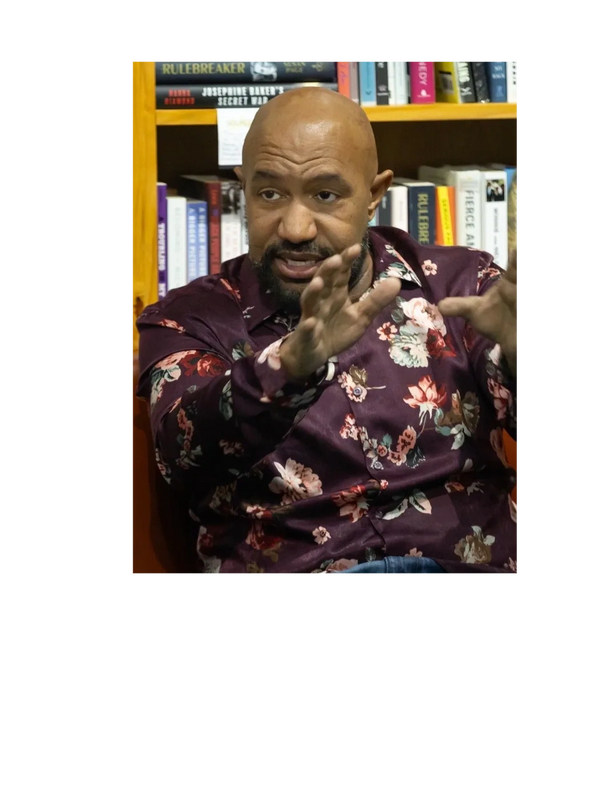 Man in floral shirt gesturing while speaking in front of a bookshelf.