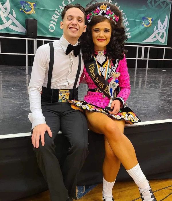 A smiling couple in Irish dance attire posing with a trophy on stage.