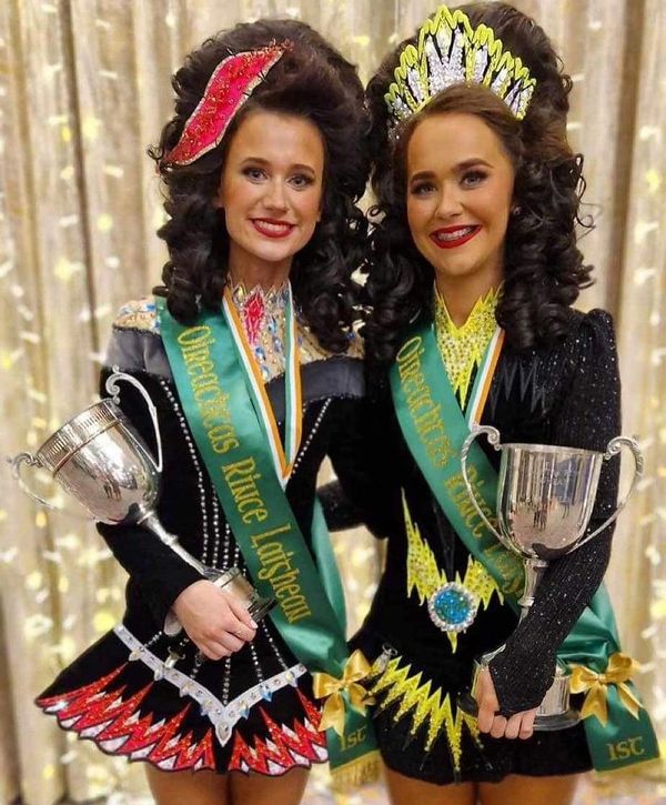 Two girls in Irish dance costumes holding trophies and wearing sashes and crowns.