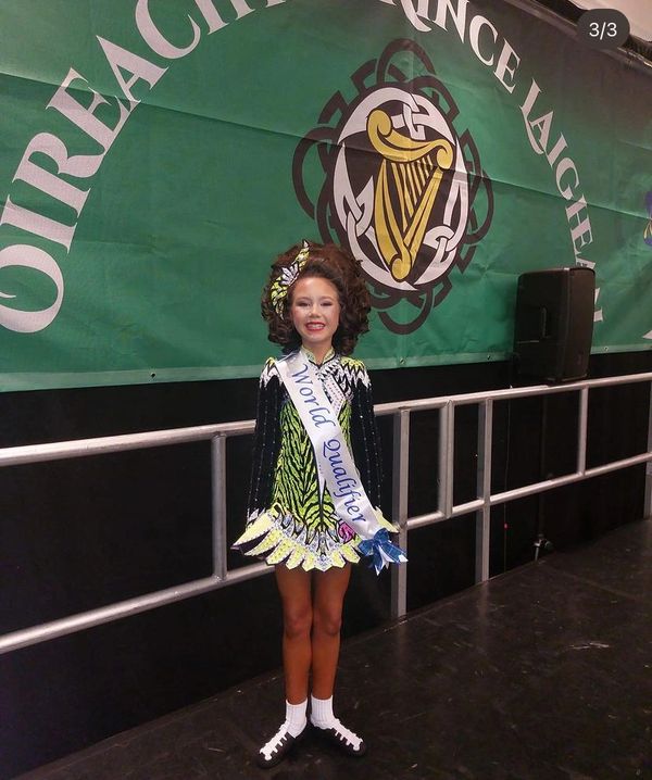 Young Irish dancer in colorful costume with 'World Qualifier' sash smiles on stage.