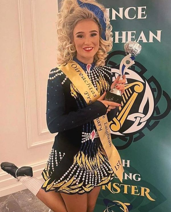 Young woman in Irish dance costume holding a trophy and wearing a winner's sash.