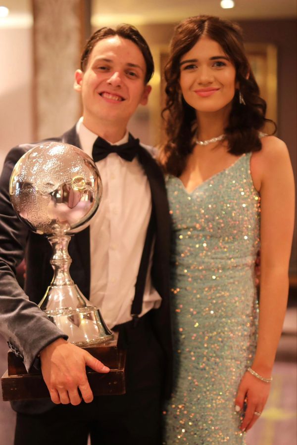 Young man in tuxedo holding a large trophy, standing with a woman in a sparkling dress.