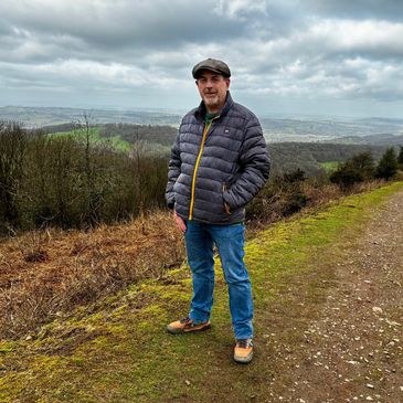 Man in outdoor jacket and cap standing on a hiking trail with a scenic landscape behind.