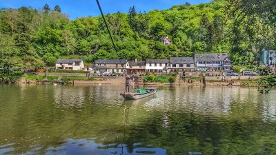 A boat crossing a river by a cable in a green, hilly village setting.