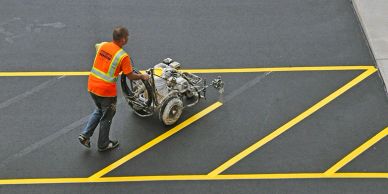 Worker in orange vest pushing a machine on asphalt with yellow lines.