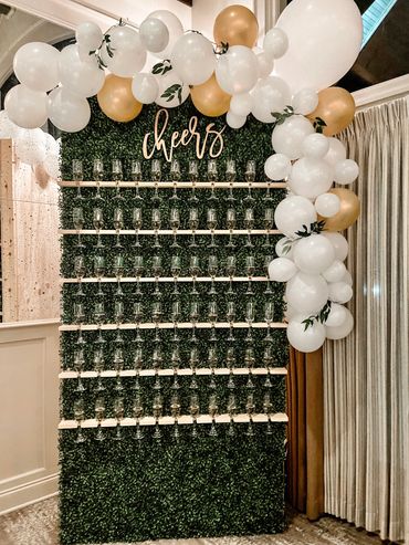 Image is of a green champagne wall with 6 rows of champagne glasses hanging on it. 