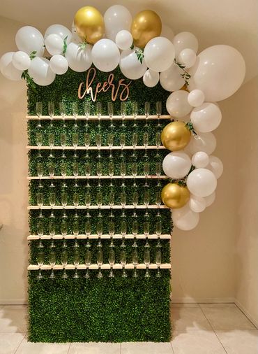 Image is of a green champagne wall with 6 rows of champagne glasses hanging on it with balloons arou