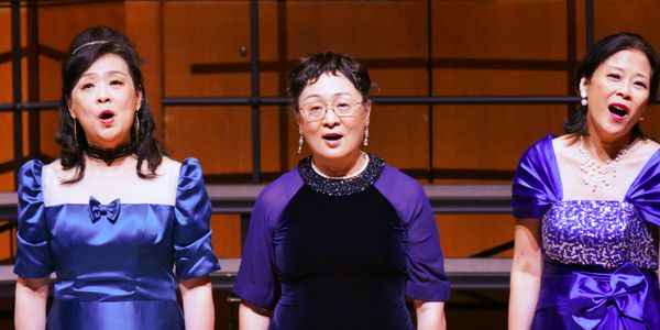Three women in elegant dresses singing on stage.