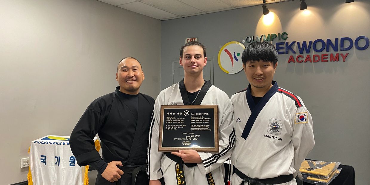 Three Taekwondo practitioners with a certification plaque inside a martial arts academy.