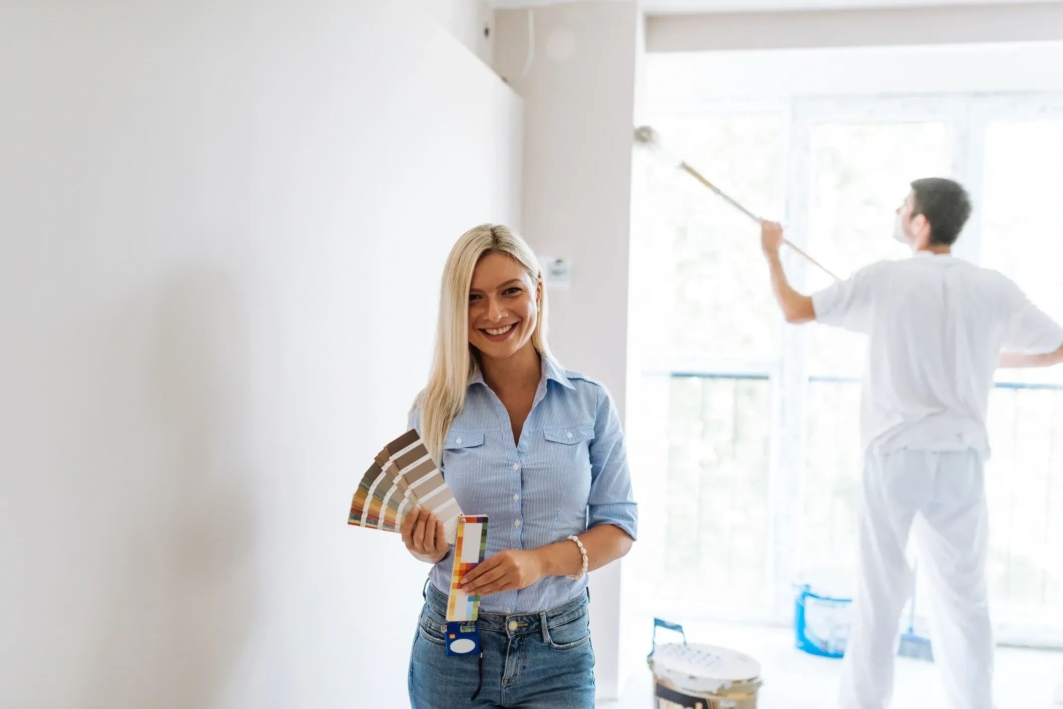 A smiling woman holding paint swatches with a man painting a wall in the background.