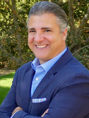 A smiling man in a blue suit jacket outdoors with arms crossed.
