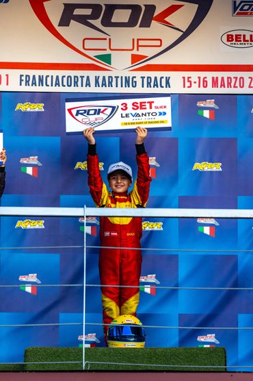 Young kart racer celebrates victory holding a prize sign on the podium.