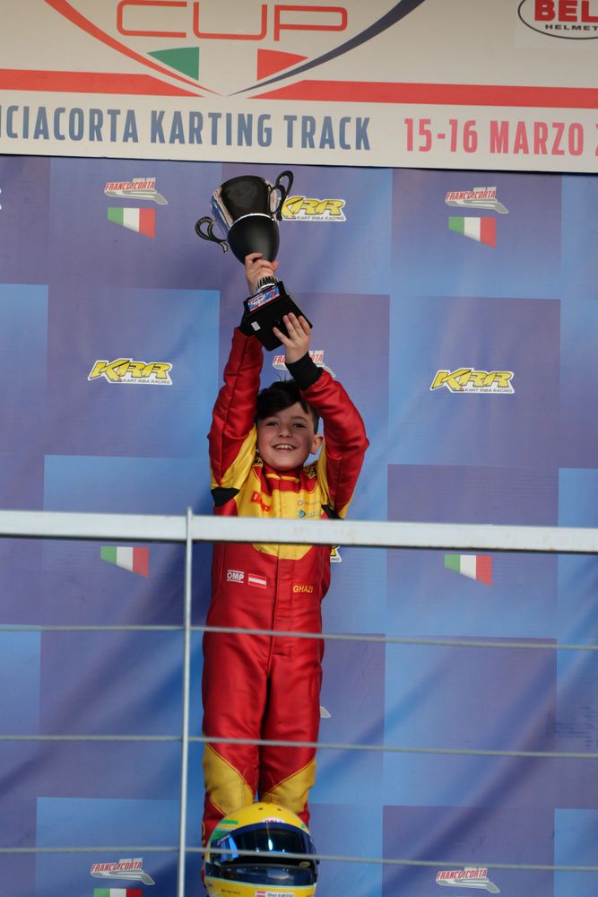 Young kart racer celebrates victory holding up a trophy at Franciacorta Karting Track.