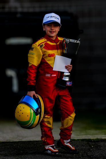 Young kart racer in red and yellow suit holding trophy and helmet.