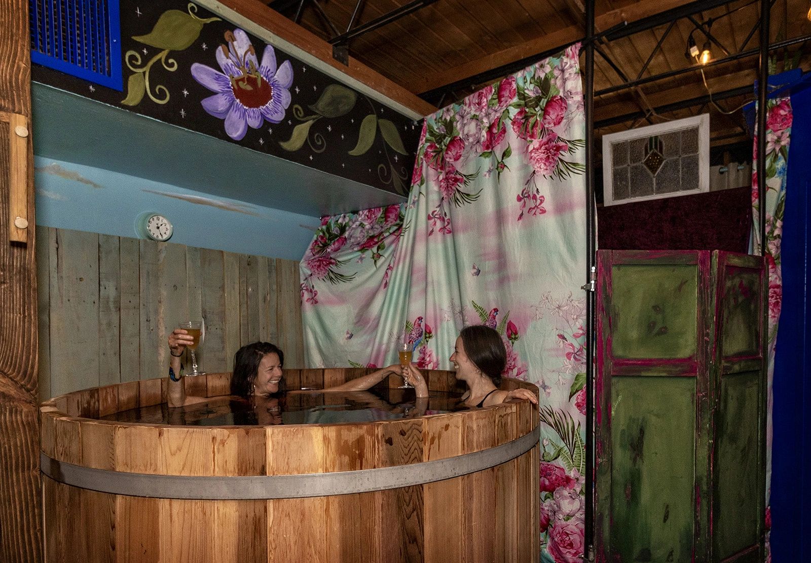 Two women enjoying drinks in a wooden hot tub indoors with floral curtains.