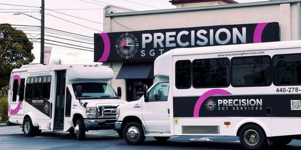 Two white buses with Precision SGT Services branding parked near a building with a matching sign.