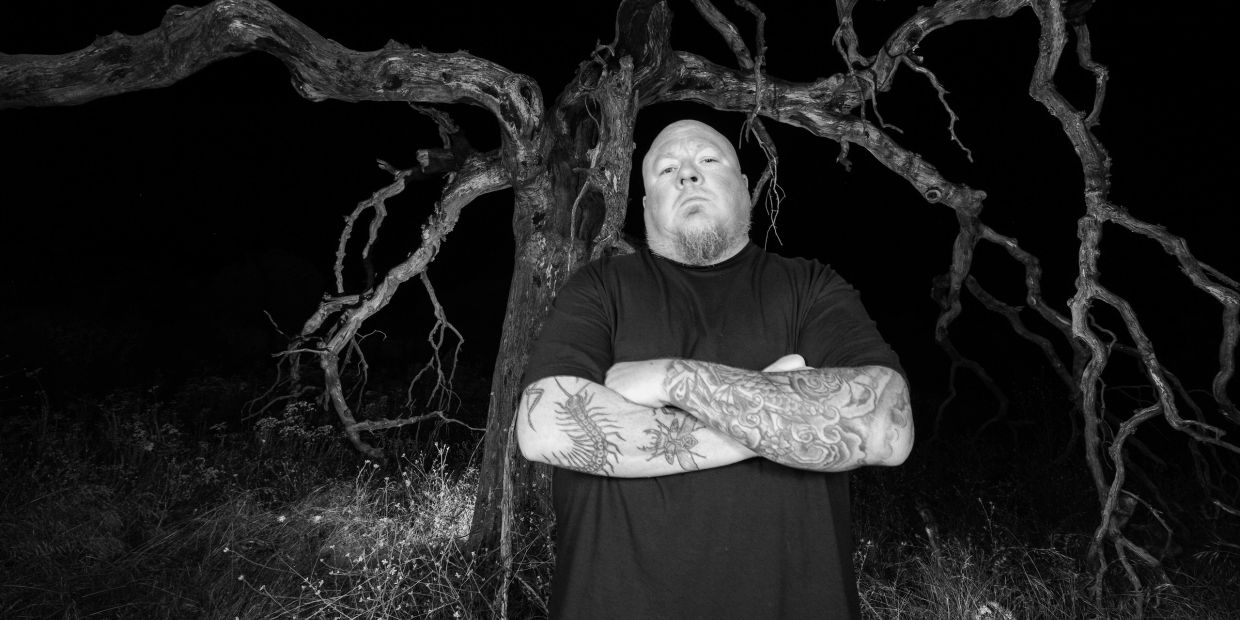 Tattooed man stands with arms crossed in front of twisted tree at night.