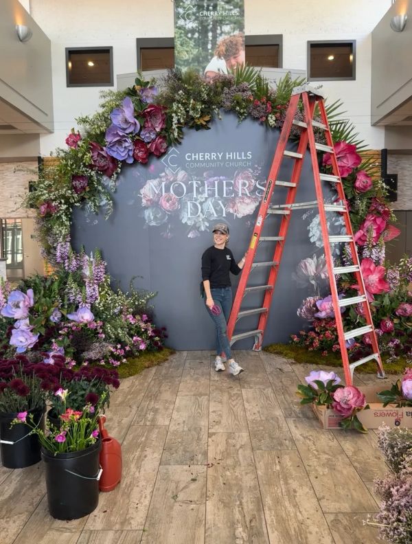 Woman standing beside a floral arch with a Mother's Day sign and a ladder.