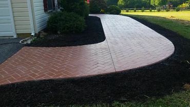 Curved brick-patterned concrete walkway bordered by dark mulch around a house.