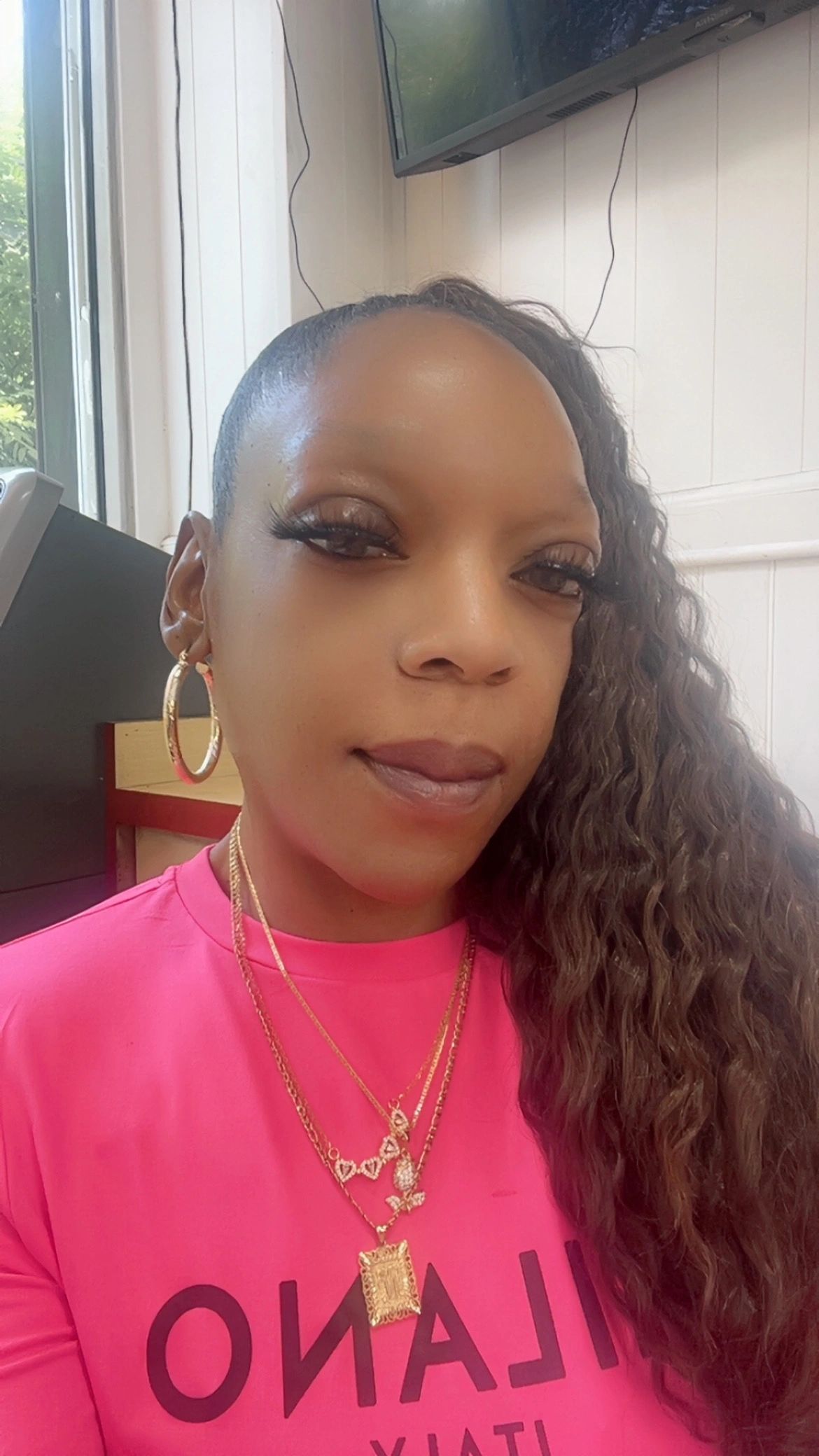Woman with long curly hair wearing gold jewelry and a pink shirt.