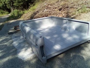 A primer-coated truck bed shell outdoors on gravel near brush.