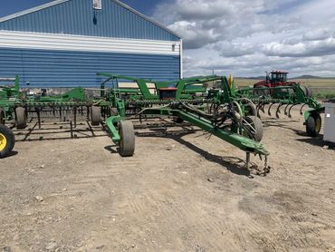 Large green farming cultivator parked on a dirt lot next to a blue and white building.