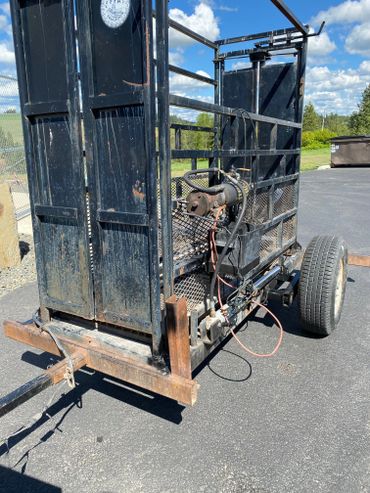 Rusty black metal trailer with mechanical parts attached, parked on asphalt under blue sky.