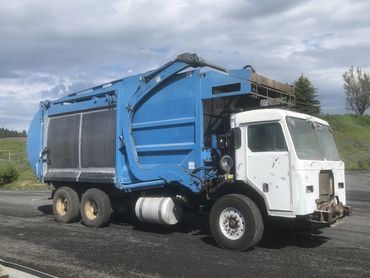 A worn blue and white garbage truck parked on asphalt under a cloudy sky.