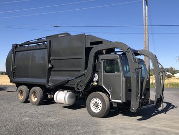 Black garbage truck with front-loading arms parked on a gravel surface.