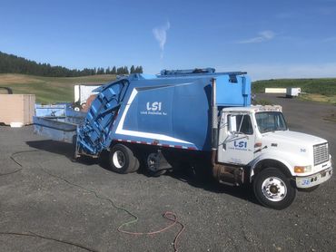 A blue and white LSI garbage truck parked outdoors on a sunny day.
