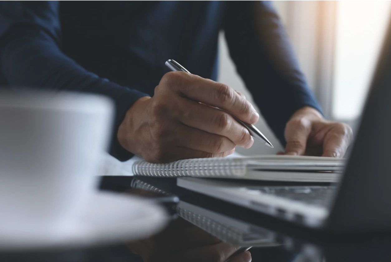 Person writing in a notebook next to a laptop and coffee cup.