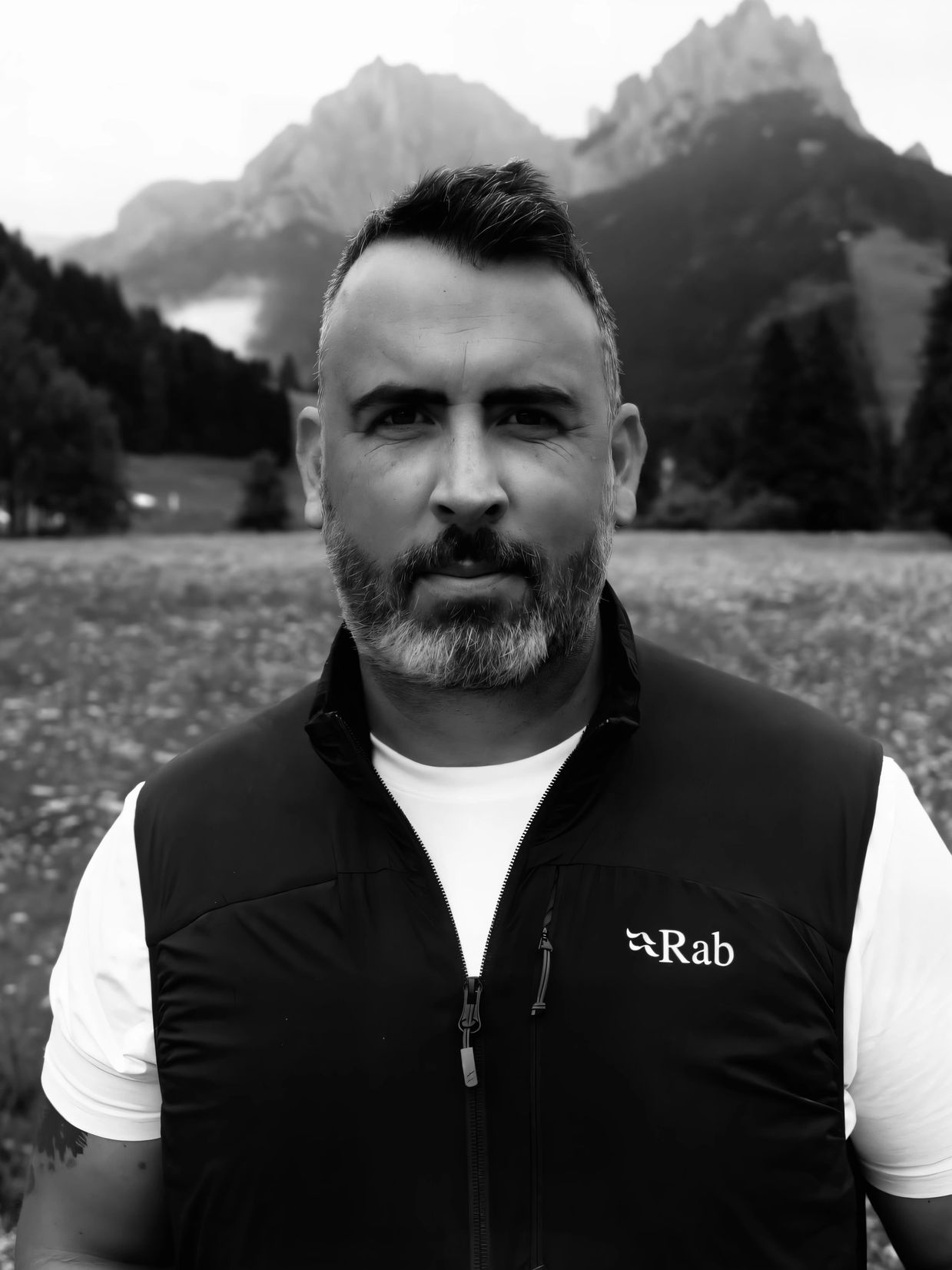 Man with a beard in outdoor vest stands in front of mountainous landscape.