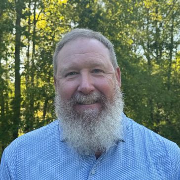 Smiling man with a full beard in a patterned shirt outdoors.