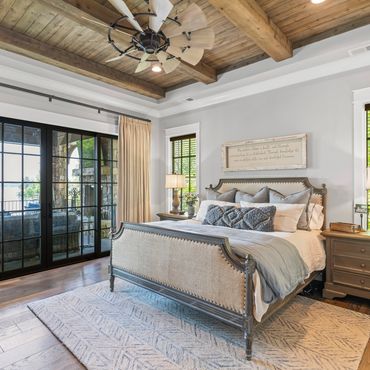 Elegant bedroom with wooden ceiling, large bed, and glass doors overlooking a patio and water view.