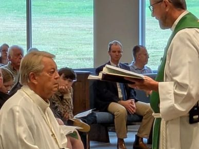 A priest reading from a book to a seated clergyman during a church service.