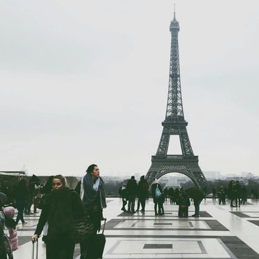 Tourists walking with luggage near the Eiffel Tower on a cloudy day.