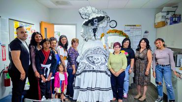 Group poses with a tall figure in elaborate Day of the Dead costume indoors.