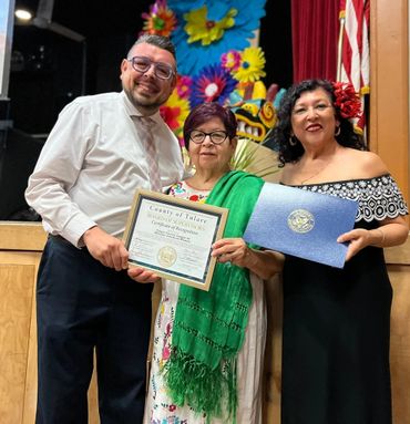 Three people smiling, holding a certificate of recognition, with colorful decorations behind them.