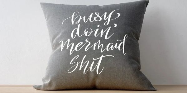 Gray pillow with white cursive text about being busy doing mermaid activities.