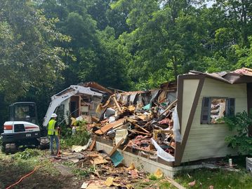 Excavator demolishing a mobile home surrounded by greenery and debris.