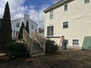 Backyard with stairs, bushes, and a ladder near a yellow house under a partly cloudy sky.