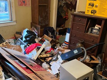 Cluttered room filled with boxes, papers, and furniture in disarray.