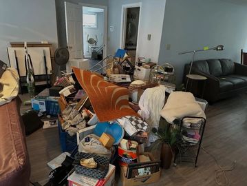 Cluttered living room with piled-up household items and furniture.